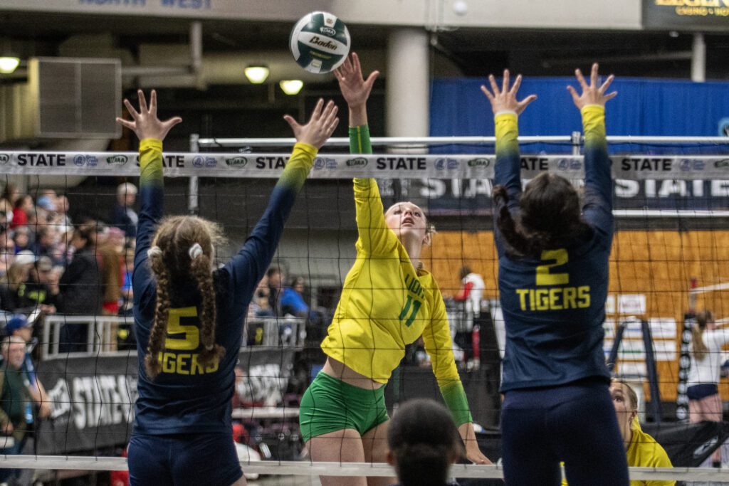 Lynden's Rian Stephan spikes over the net as two defenders leap into the air with both their arms up to block the shot.
