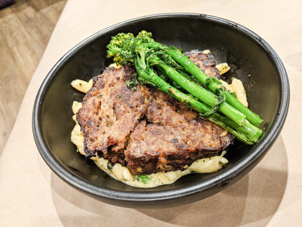 A black bowl of pasta topped with meatloaf and broccolini.