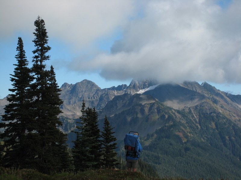 The scenic view of the Pacific Northwest Trail as a person wearing a hiking backpack stands to admire the mountains where the peaks are covered with clouds overhead.