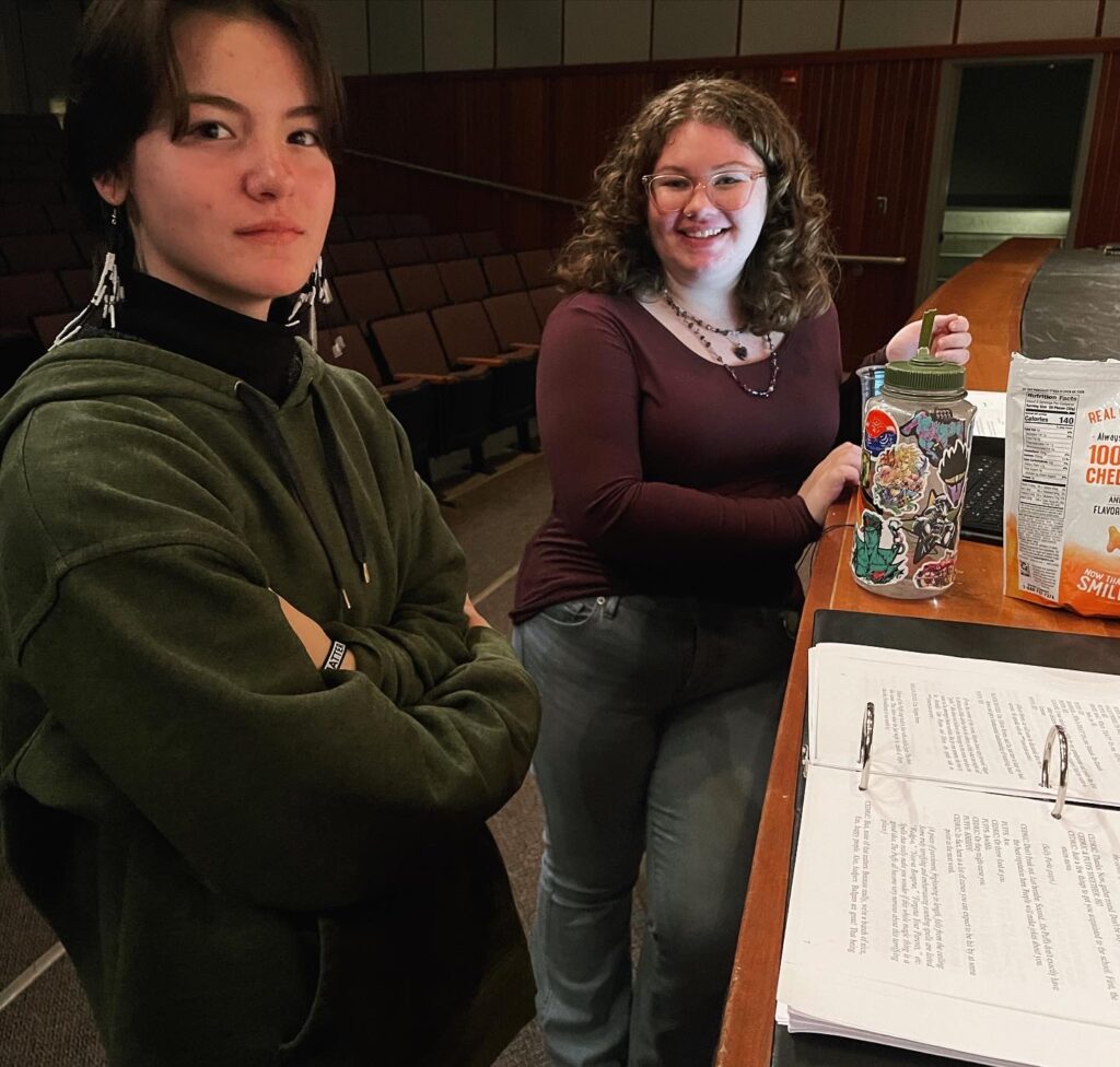 Stage manager Lauren Mahoney (right) and assistant stage manager Teoxihuitl Larsen smile for the camera next to the stage where they have kept their personal belongings along with the script binder.