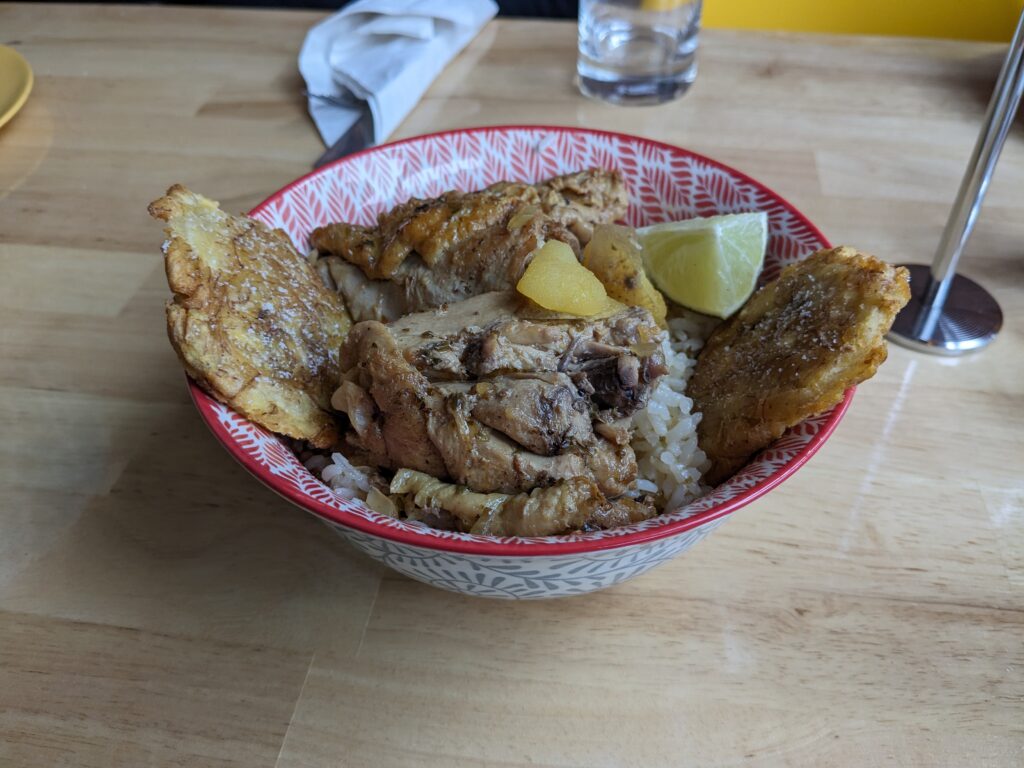 Pollo guisado, a bowl of white rice and tostones topped with tender, bone-in, stewed chicken and veggies, is served in a decorated red and white bowl.