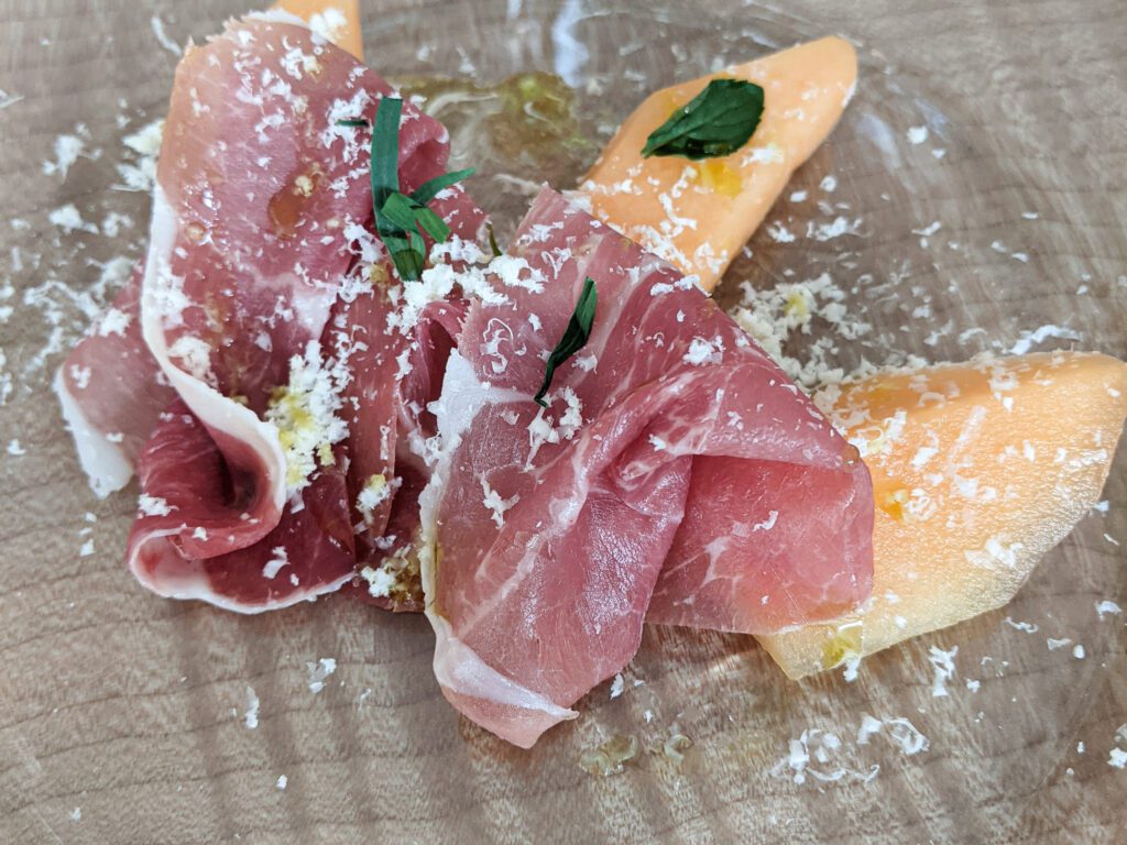 Prosciutto, carbonated melon, and garnished with white shavings and green herbs are served on a clear plate.