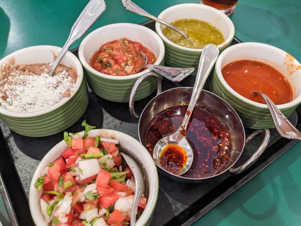 A tray full of dips and salsas is served with spoons and on a basket of whole fried small tortillas.