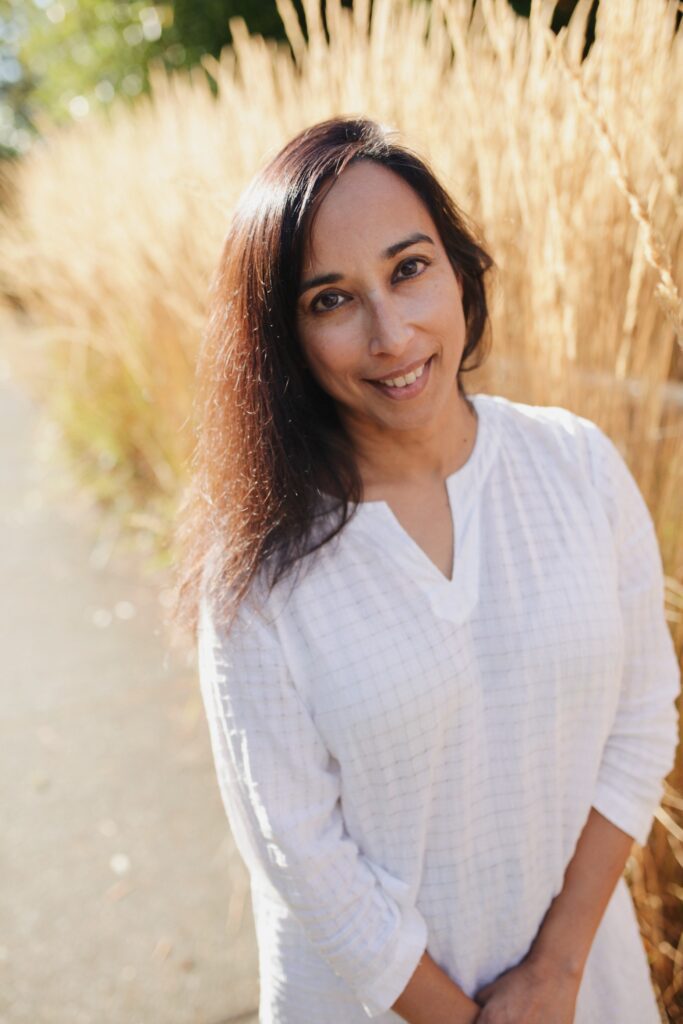 Sati Mookherjee, author of "Ways of Being", smiles for the camera next to tall grass.