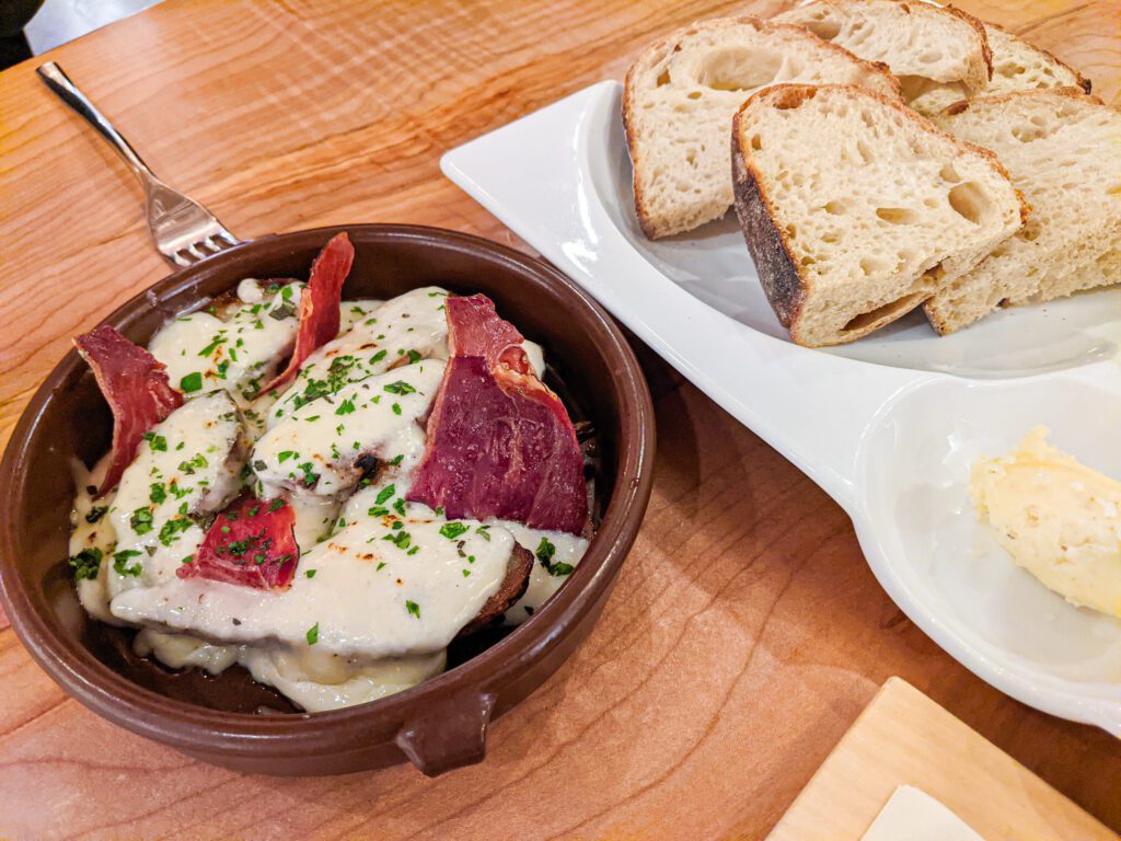 Roasted sunchokes drenched in a béchamel sauce, gruyere cheese and prosciutto next to a bread platter.