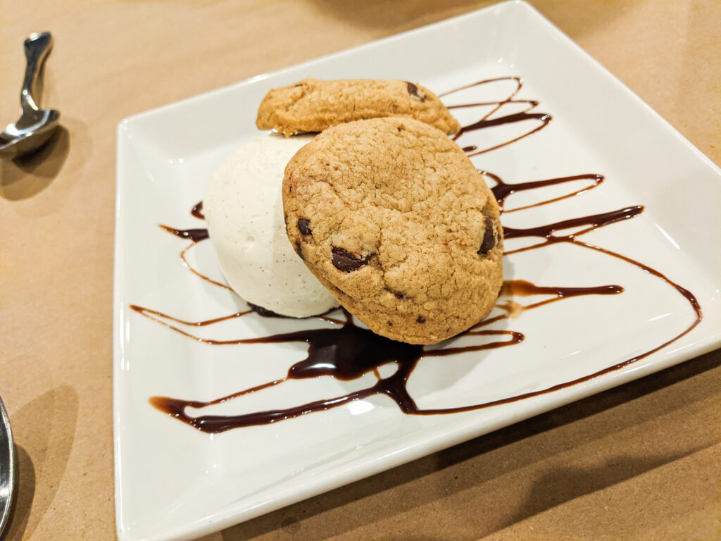 Two chocolate chip cookies are stacked on top of vanilla ice cream drizzled with chocolate sauce.