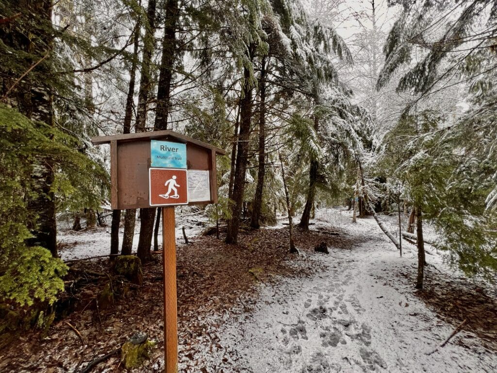 The River snowshoe route's path is covered in snow as a small information board shows the trail map.