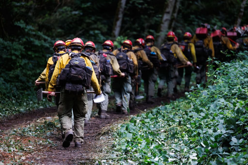 Firefighting crews from the Baker River Hotshots head to their trucks in a single file line, all wearing safety gear and helmets.