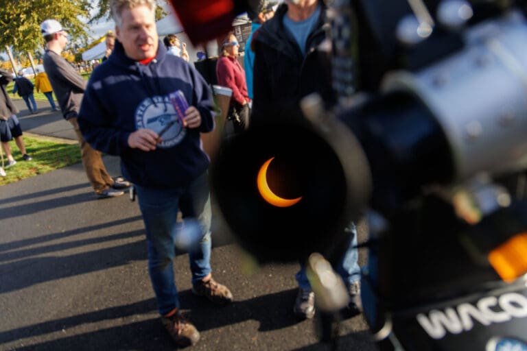 As seen through a camera, the moon covers the sun partially to show a crescent shape as attendees chat around the telescope.