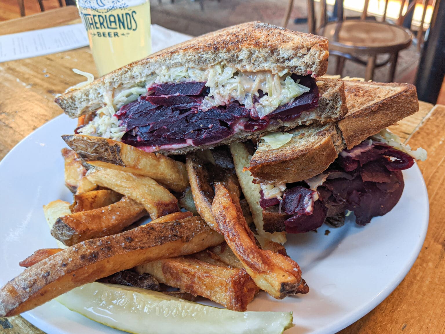 A plate of beet Reuben consists of marbled rye bread, sauerkraut, swiss cheese, and Russian dressing stacked next to fries.