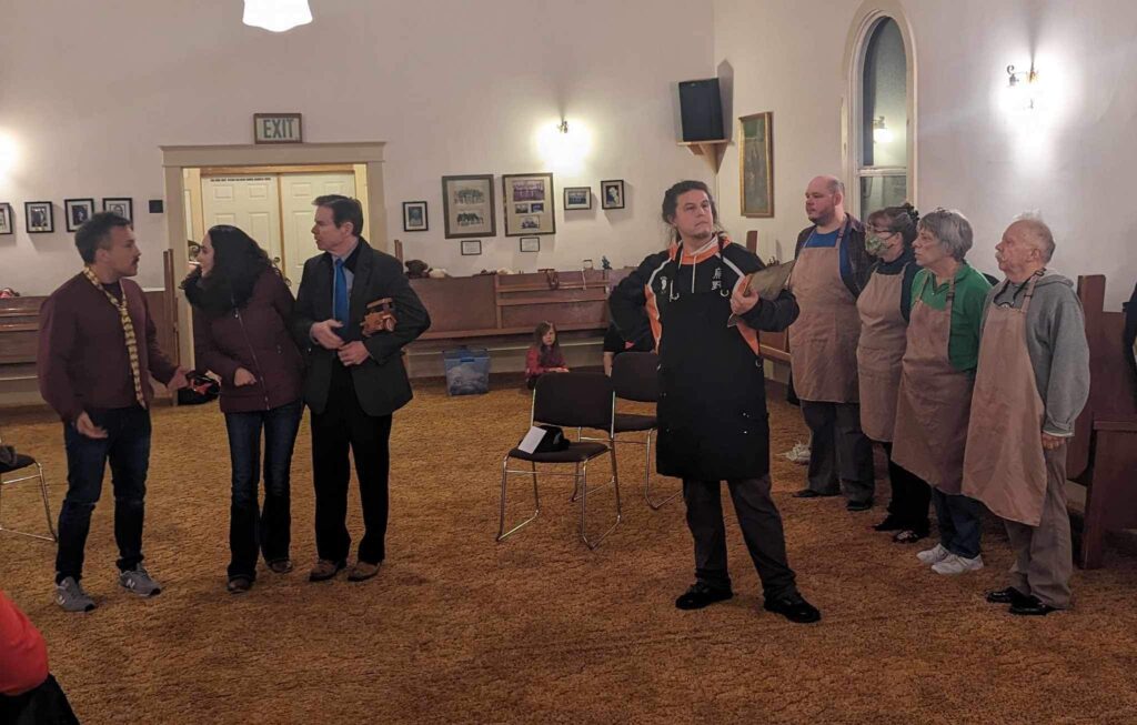 Actor Sandy Brewer, far right, joins cast mates for a rehearsal of "The Man Who Saved Christmas," who are lined up wearing brown aprons as attendees chat to a staff member.