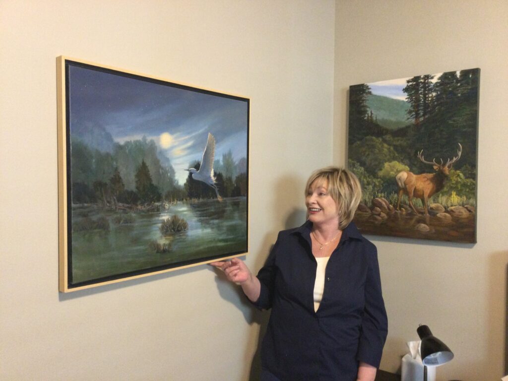 Sheila Steinborn stands in between two paintings as she points to the one on the left.