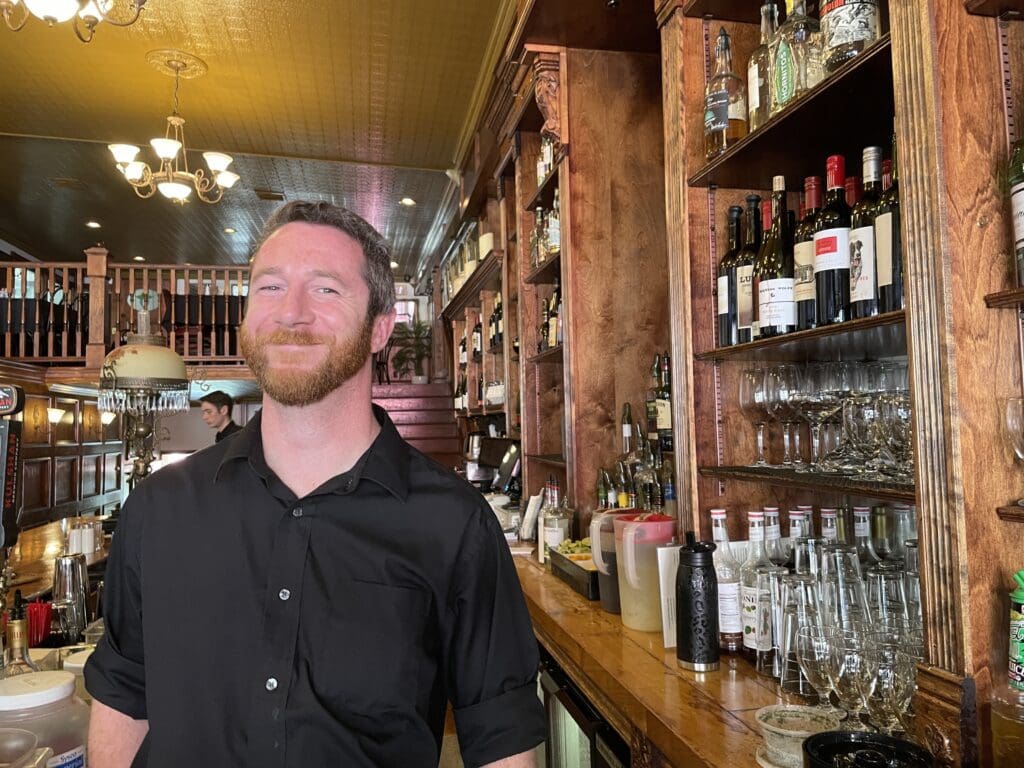 John Drum poses at the bar at Skylark's Hidden Cafe as he smiles for the camera next to shelves filled with different assortments of drinks.