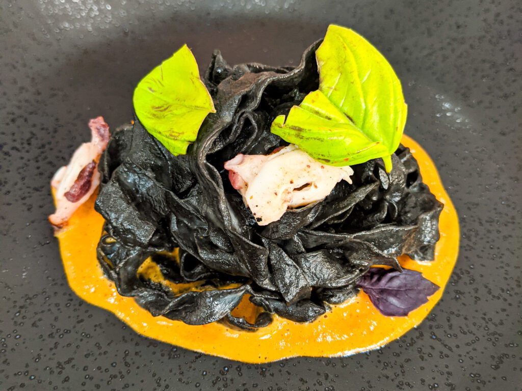Delicate squid ink pasta with charred octopus and bright orange chorizo foam served with green herbs as garnish and placed on a black plate.