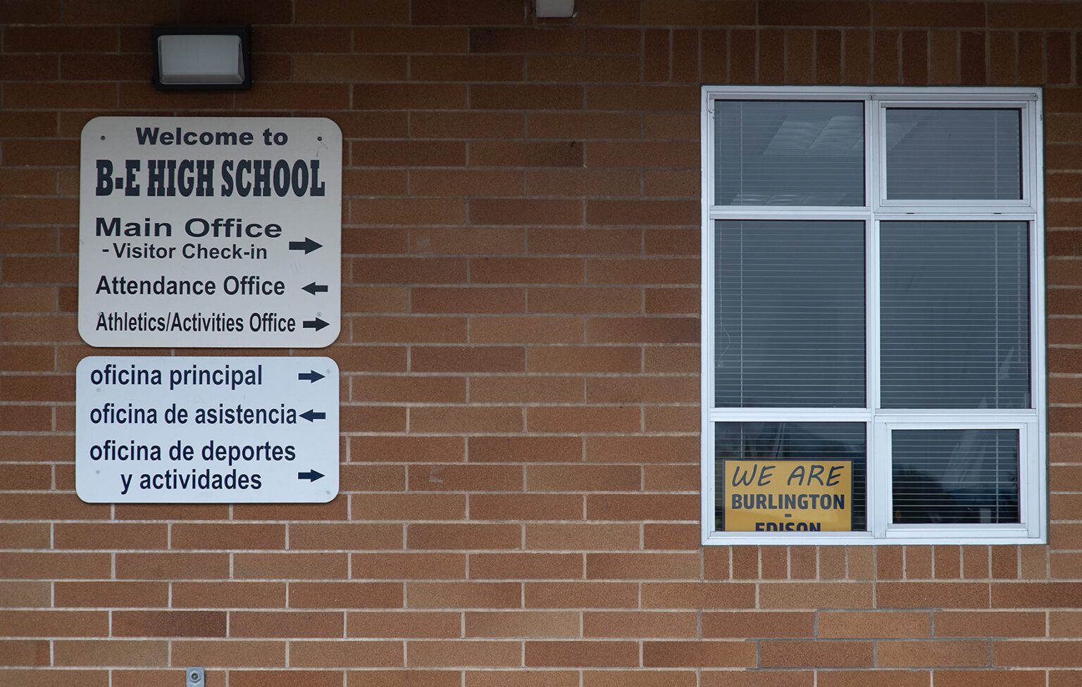Directional signs on a brick wall are placed next to a window with a yellow sign that reads " We are Burlington - Edison".