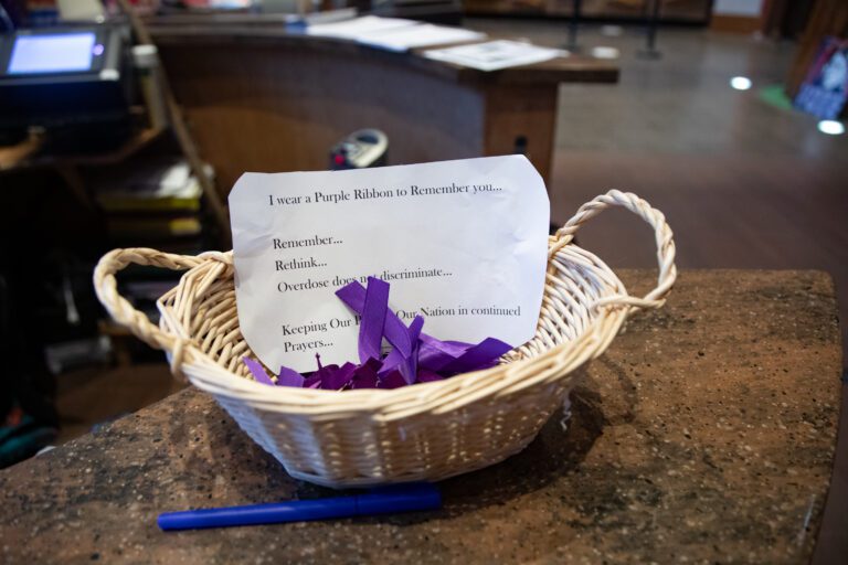 A woven basket next to a blue pen holds purple ribbons and a note to raise awareness about overdosing.