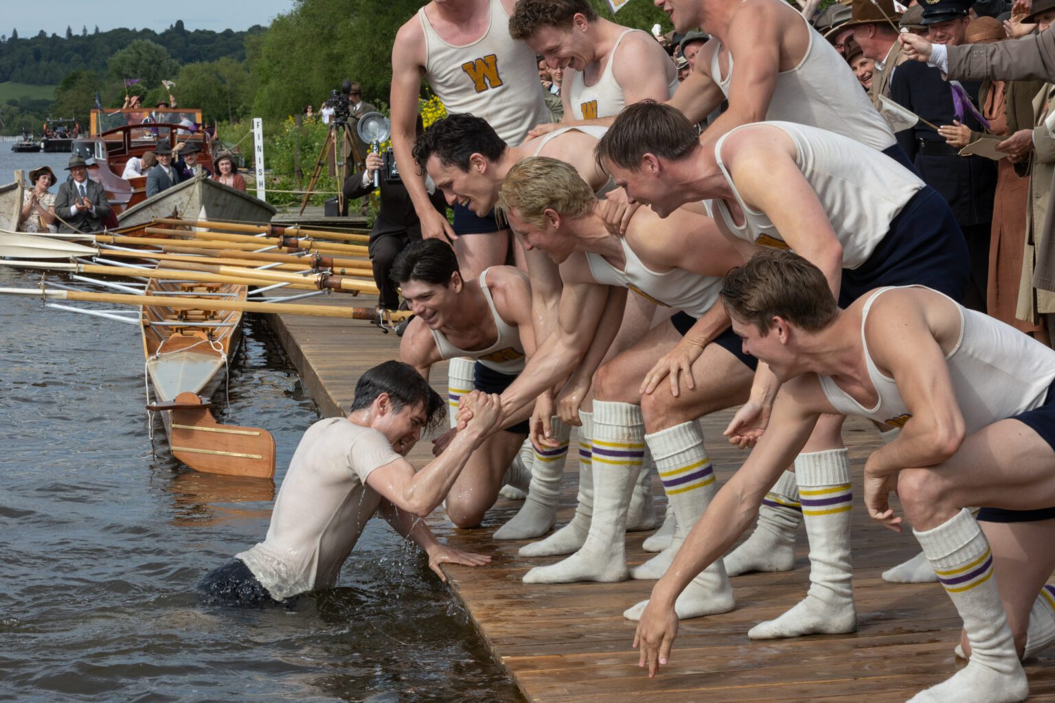In a movie scene, young men in tank tops and socks pull a rowing teammate out of the lake next to a row boat.