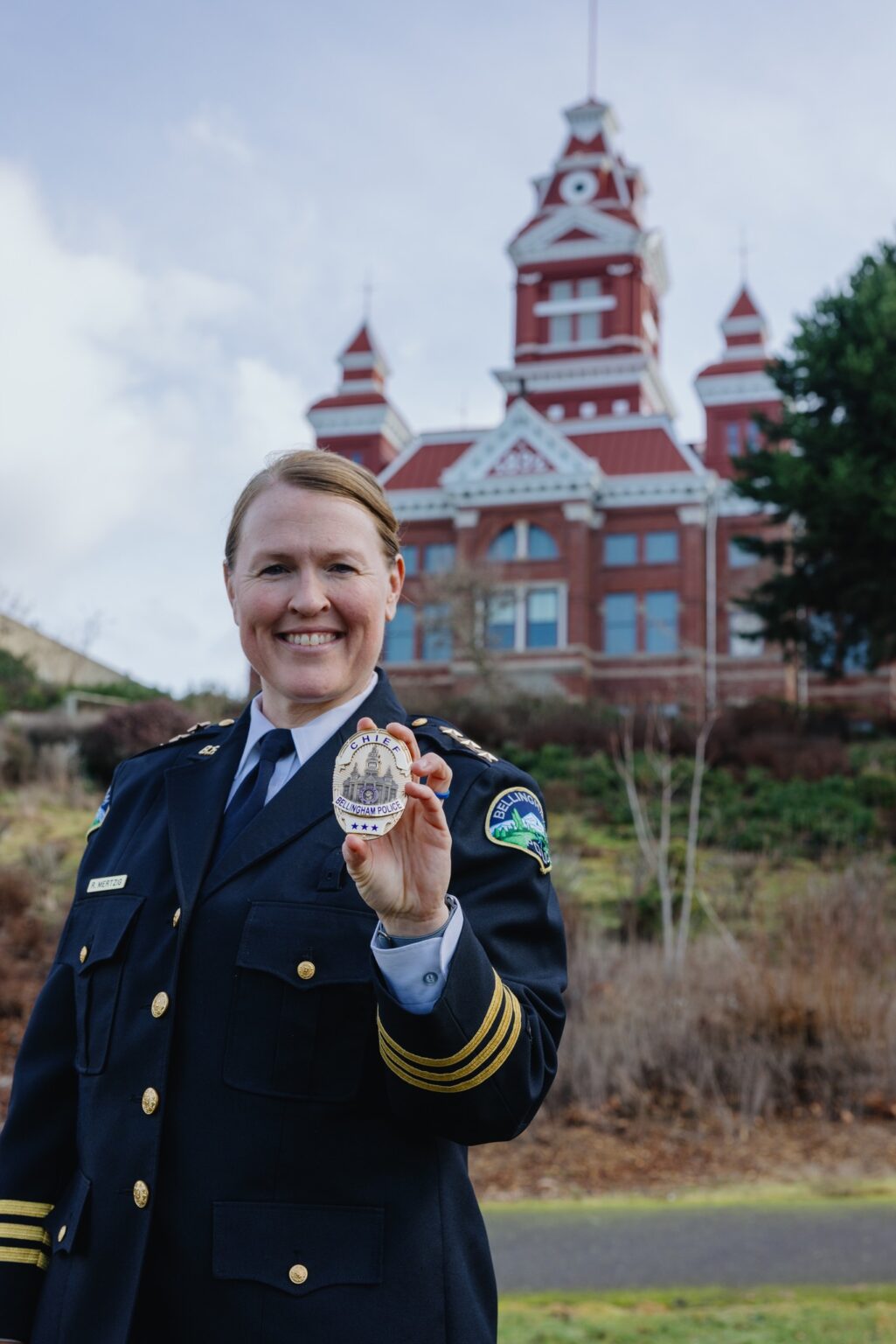 A woman with blond hair pulled back in a navy police uniform holds a police badge in front of a red building.