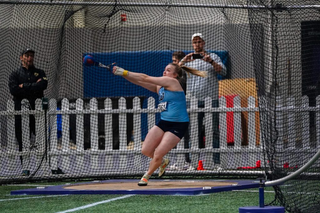 Western Washington University's Katie Potts loads up a throw during the weight throw event as a spectator takes a photo from behind the black net and white fences.