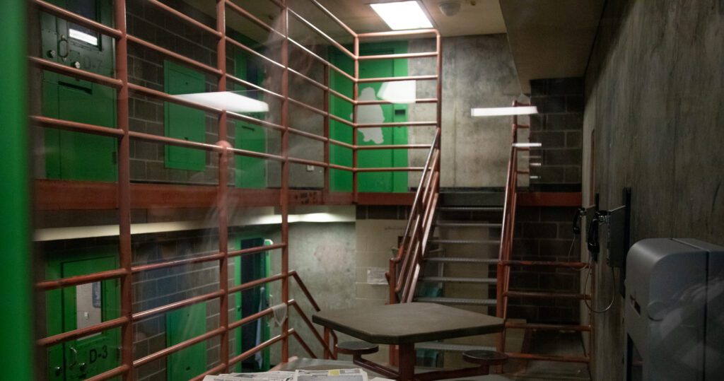A photo through the window looking into where inmates are kept. There are phones on the walls on the right, a table with chairs in the center, and red stairways that lead to the green doors hosting cells.