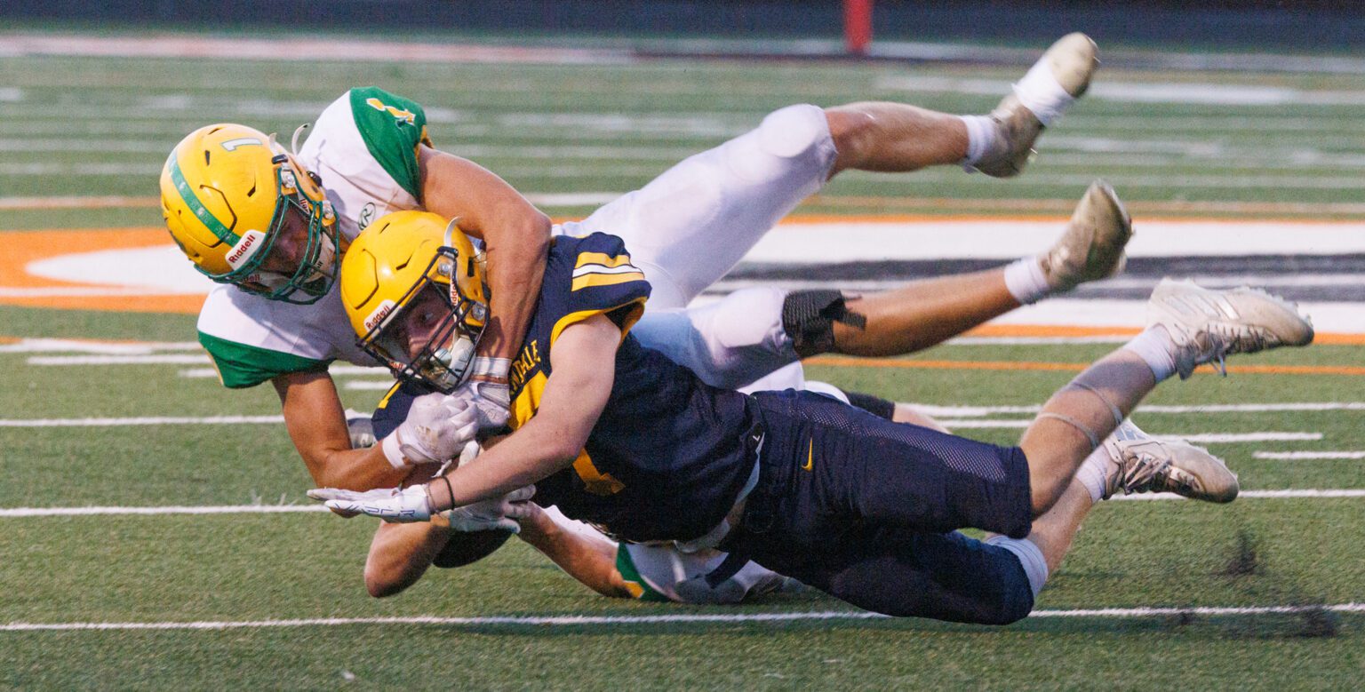 Zach Neilsen holding onto the football to his chest as he is tackled by Kobe Baar and one other player.