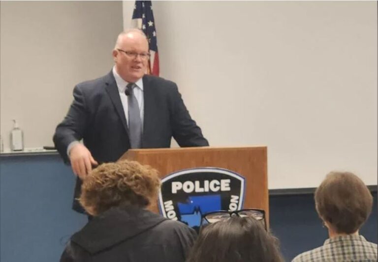 A white man with glasses and a black suit, speaks at a brown podium with a sign that reads Police: Mount Vernon.