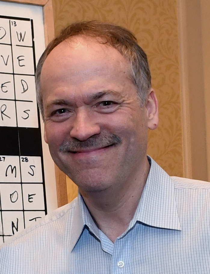 Will Shortz smiling for the photo in front of a finished crossword board.