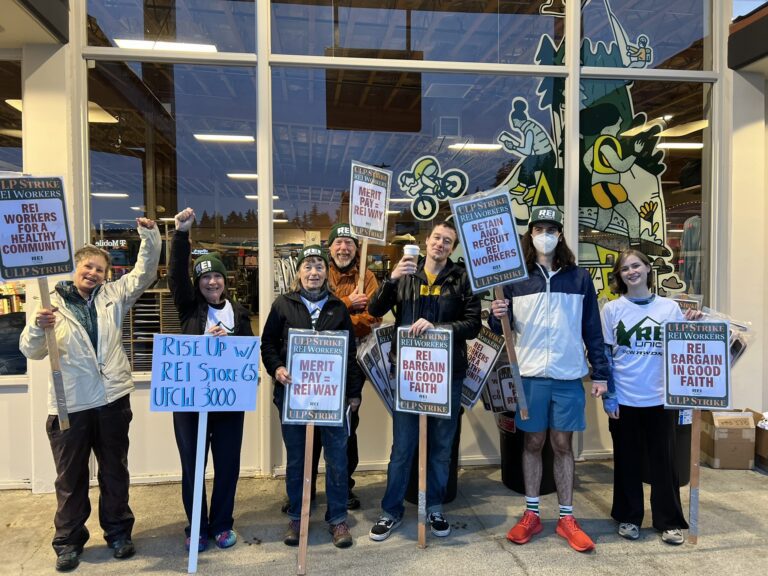People in jackets and hats holding signs in front of the REI store in Bellingham.