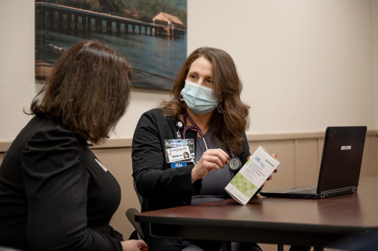 A woman with brown hair with a stethoscope around her neck and wearing a blue mask, holds a white box while looking at a woman with brown hair.