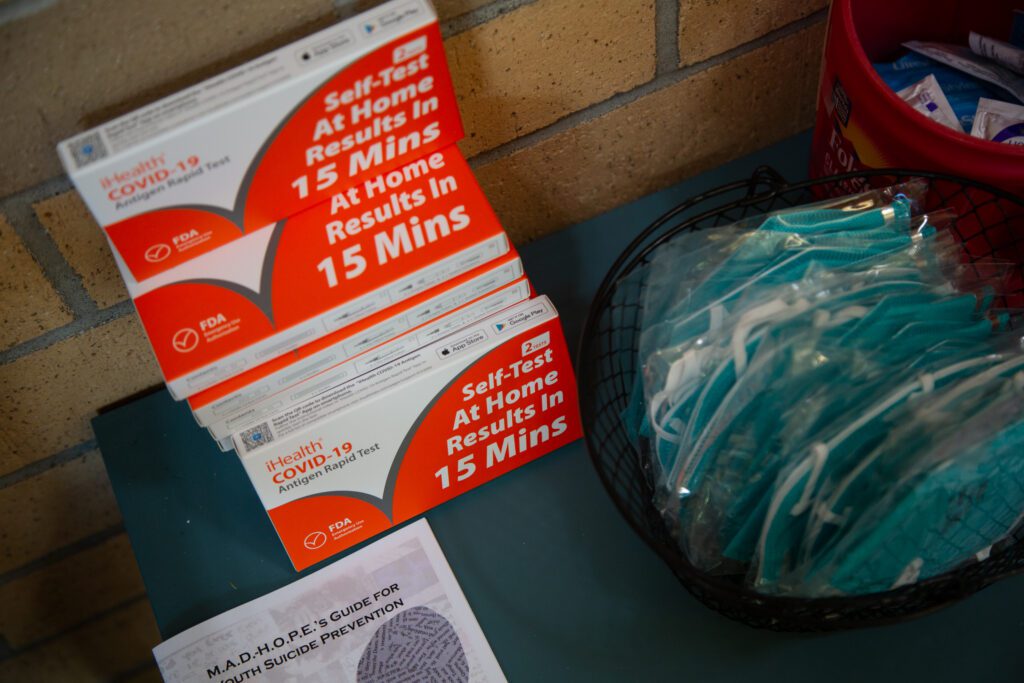 Masks and COVID-19 tests on a table next to hand wipes and a guide for youth suicide prevention.