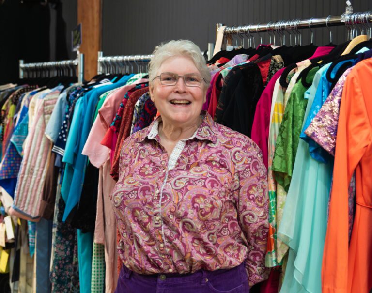 Genny Cohn stands in front of rows of clothes of varying colors and fabrics.